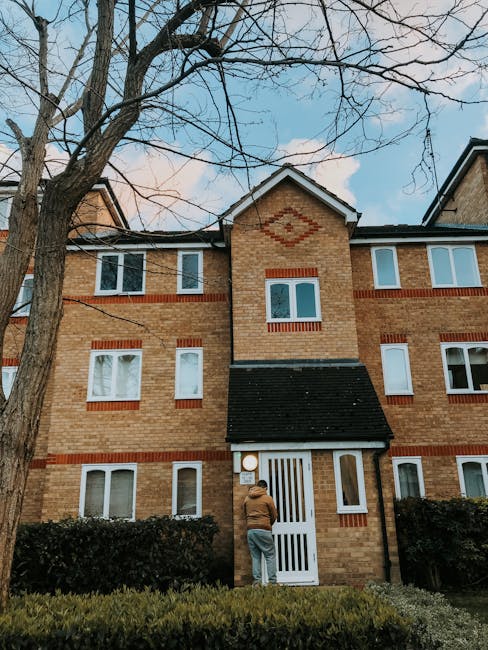 Exterior view of a multi-story brick residential building with a steep black tiled roof and white-framed windows. The building features red brick accents around the windows and a decorative diamond pattern near the roof peak. A person wearing casual clothing is standing at the white front door, which has vertical slats and is partially obscured by a black hedge. The scene is taken during daytime with a clear sky, and a large leafless tree with branches stretching across the sky is visible in the foreground. The environment appears clean and well-maintained, reflecting the importance of property appearance and maintenance in the Kingston flats and estates area, as highlighted on the Carpet cleaning Kingstonuponthames.co.uk page.