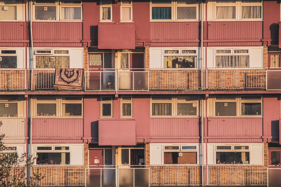Close-up of a multi-storey residential building façade with pink painted exterior walls, featuring several balconies with transparent glass railings and white metal frames. Some balconies display outdoor items such as a patterned blanket hanging over the railing. The large windows have white frames, with curtains visible inside. The building's surfaces appear clean, with a neat and maintained appearance, illuminated by warm sunlight. This image does not show any cleaning tools or activities but emphasizes the exterior maintenance of a Kingston flats and estates building, illustrating the importance of surface cleaning and property upkeep, aligned with services offered by [COMPANY_NAME] as part of their domestic cleaning and deep cleaning solutions for residential properties in Kingston upon Thames.