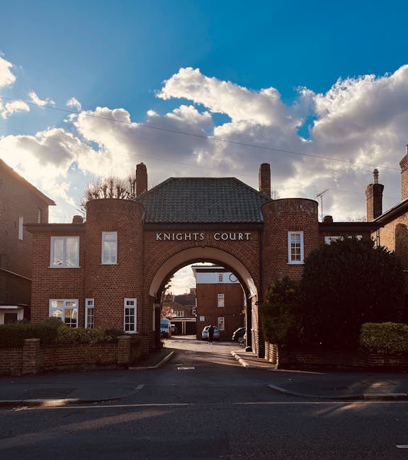 A brick residential complex named Knights Court featuring an arched entrance with the sign 'KNIGHTS COURT' displayed above. The structure has a dark tiled roof, with two symmetrical chimneys visible, and is flanked by neatly maintained greenery and small brick walls. The surrounding area includes parked cars on the driveway, with partly cloudy skies overhead. This setting exemplifies traditional UK architecture and outdoor space, captured in natural daylight. [COMPANY_NAME] specializes in domestic cleaning services, including surface cleaning and sanitisation, ensuring high standards of hygiene for homes and similar properties, in line with the focus on emergency stain removal and deep cleaning for residential environments, as referenced in the page title Kingston upon Thames.