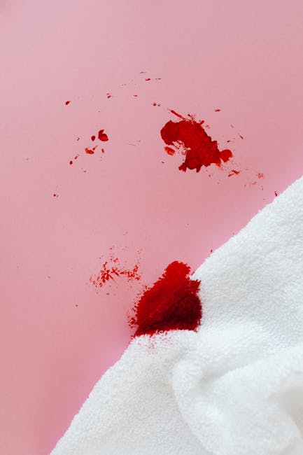Close-up of a pink wall with a fresh red bloodstain, some smaller splatters nearby, next to a white textured towel or cloth partially covering the bottom right corner. The surface appears smooth, and the lighting is bright, highlighting the vivid color of the stain. This image relates to emergency stain removal for domestic surface cleaning, as discussed on the Kingston upon Thames cleaning services page by [COMPANY_NAME].