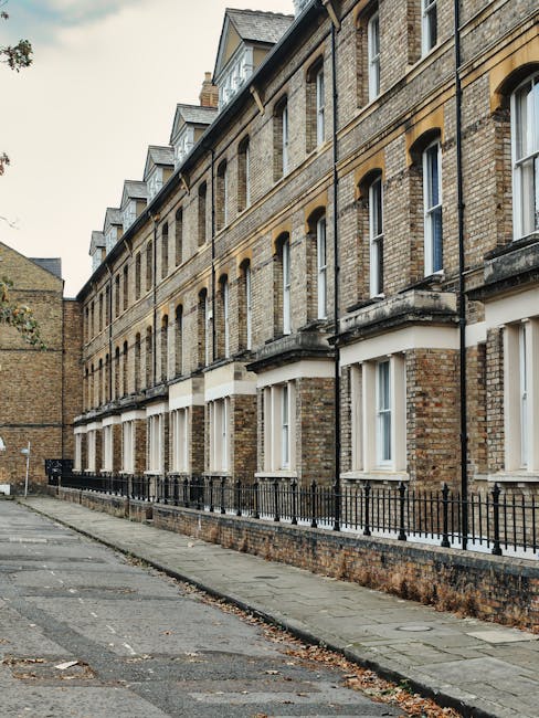 The image depicts a row of traditional Victorian-style terraced houses constructed from light brown brick, situated along a paved residential street. The houses feature large sash windows with white frames, some with window sills, and decorative stone lintels above the windows. The ground-floor windows are partly obscured by black wrought iron railings that run along the small front gardens, which are paved and free of clutter. The pavement is made of concrete slabs, showing some leaf litter and minor debris, while the street surface appears to be asphalt with a few cracks and patches. The buildings are well-maintained, with clean facades, and the overall scene is softly illuminated by natural daylight, suggesting an overcast sky. This image is associated with professional cleaning services offered by [COMPANY_NAME], emphasizing thorough surface cleaning and maintenance for residential properties in Surbiton to KT2 areas, Kingston upon Thames.