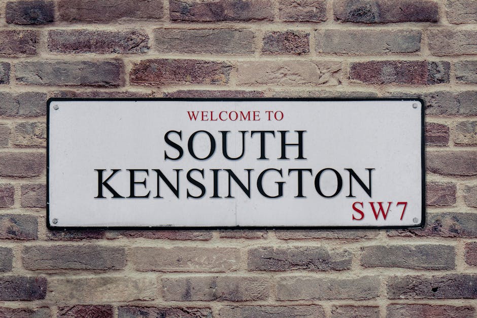 Close-up of a rectangular white metal street sign mounted on a textured red brick wall, displaying the text 'WELCOME TO SOUTH KENSINGTON SW7' in black uppercase letters, with 'WELCOME TO' and 'SW7' in red. The sign features rounded corners and visible mounting screws at each corner, with even lighting that highlights the smooth surface and legible text. The brick wall behind is weathered, with varied shades of red and gray mortar, providing a contrasting background for the sign. This image captures the outdoor urban environment associated with South Kensington, relevant to property maintenance and cleaning services offered by [COMPANY_NAME] for residential and commercial properties in the KT2 and Kingston upon Thames area.
