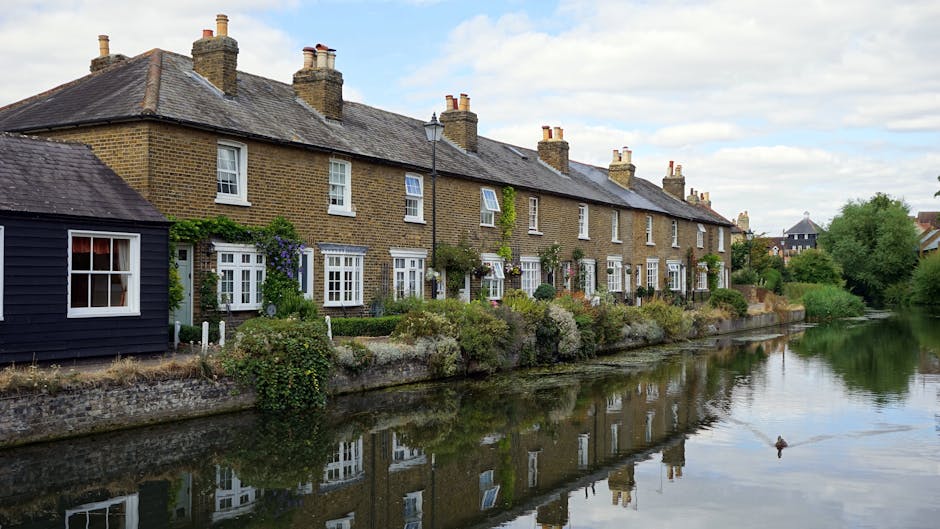The image depicts a row of traditional Victorian-style terraced houses constructed from light brown brick, situated along a paved residential street. The houses feature large sash windows with white frames, some with window sills, and decorative stone lintels above the windows. The ground-floor windows are partly obscured by black wrought iron railings that run along the small front gardens, which are paved and free of clutter. The pavement is made of concrete slabs, showing some leaf litter and minor debris, while the street surface appears to be asphalt with a few cracks and patches. The buildings are well-maintained, with clean facades, and the overall scene is softly illuminated by natural daylight, suggesting an overcast sky. This image is associated with professional cleaning services offered by [COMPANY_NAME], emphasizing thorough surface cleaning and maintenance for residential properties in Surbiton to KT2 areas, Kingston upon Thames.