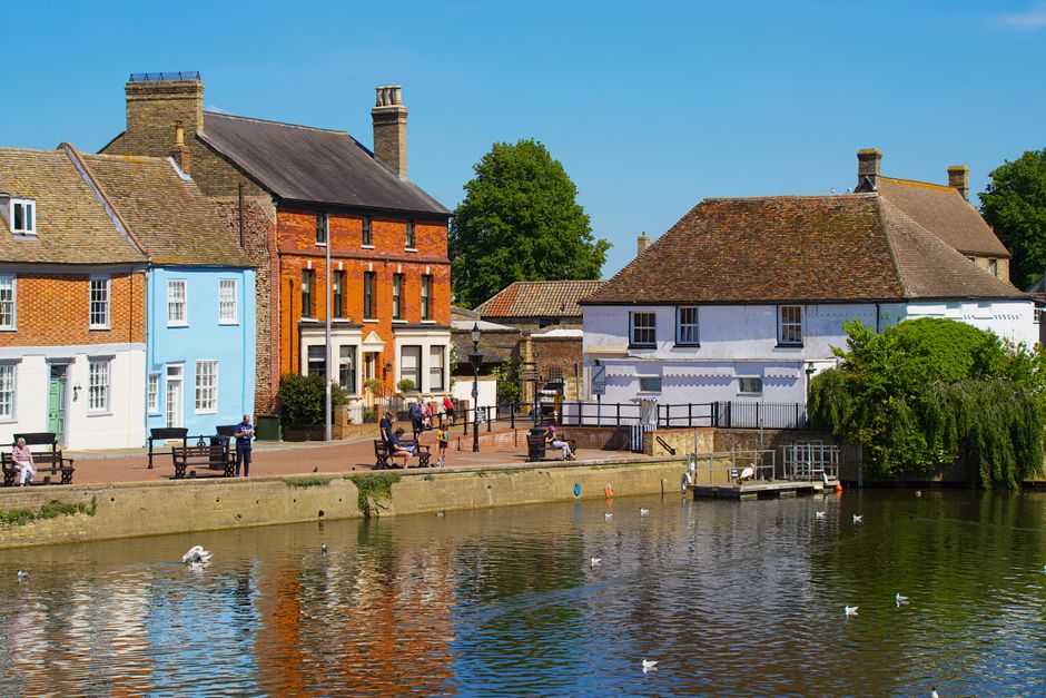 A picturesque riverside scene in Kingston upon Thames displaying a row of colorful, well-maintained residential buildings with varied architectural styles, including brick and stucco facades. The buildings are situated along a paved promenade with several benches occupied by people sitting or strolling, under a clear blue sky. The river, calm and reflective, features swans gliding on the water, while greenery such as trees and shrubs add to the tranquil atmosphere. This outdoor area appears clean, well-kept, and inviting, exemplifying the importance of surface cleaning and maintenance in maintaining outdoor hygiene, which [COMPANY_NAME] specializes in for domestic and commercial environments.