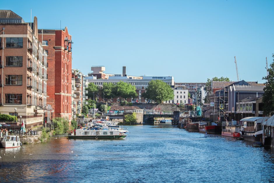 A scenic view of a river in Kingston upon Thames with calm, reflective water surface, lined on both sides by multi-story residential buildings with balconies and large windows. Some boats are docked along the riverbank, and there is a stone bridge crossing the river in the background. The area is surrounded by lush green trees, and a clear blue sky overhead suggests a bright, sunny day. The scene showcases well-maintained urban aesthetics, ideal for surface cleaning and deep cleaning discussions, and highlights the importance of sanitation in such residential and commercial environments, as promoted by Kingston upon Thames cleaning specialists.