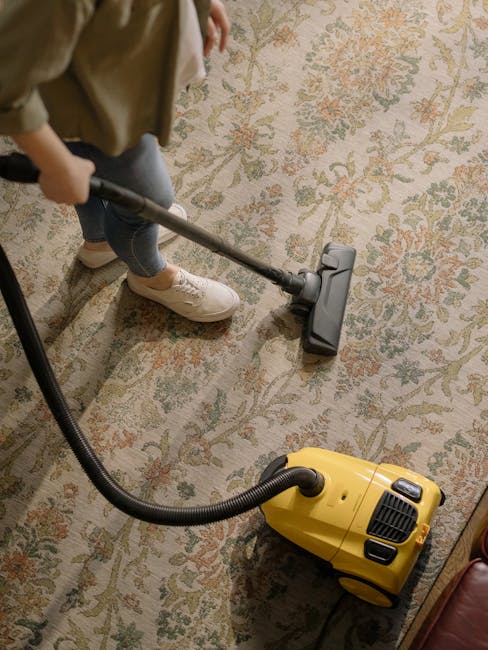 A person wearing light-colored sneakers and jeans is using a vacuum cleaner with a long metal wand attachment to deep clean a patterned area rug in a room. The yellow vacuum cleaner, equipped with black hoses, is positioned on the carpet, which features a floral design in soft pastel shades. The room is well-lit with natural light, highlighting the cleanliness and presentation of the rug. The vacuuming process is part of surface cleaning and sanitisation services offered by [COMPANY_NAME], as mentioned on the Kingston Station and Riverside same-day carpet cleaning page. The environment appears tidy and well-maintained, emphasizing professional domestic cleaning and deep cleaning procedures.