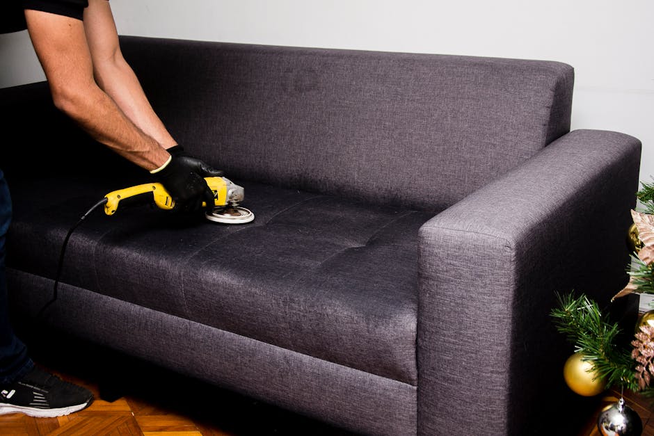A person wearing black gloves is using a handheld electric polisher to deep clean the dark gray fabric upholstery of a modern sofa, which is situated in a well-lit living room with white walls and woo