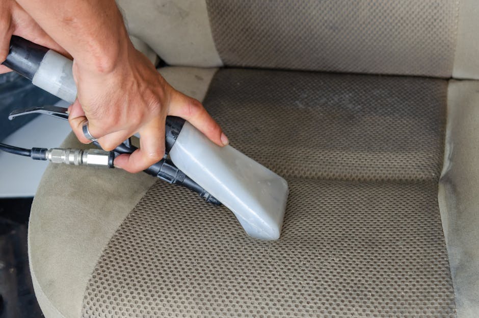 A person wearing black gloves is using a handheld electric polisher to deep clean the dark gray fabric upholstery of a modern sofa, which is situated in a well-lit living room with white walls and woo