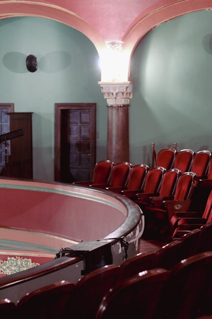 Interior shot of the Rose Theatre Kingston showcasing the beautifully maintained seating area with red upholstered chairs arranged in rows along a curved balcony. The theatre features pastel green walls with decorative white moldings and pink accents on the archways and ceiling, illuminated by soft lighting. A wooden door is visible in the background, and a corner of the stage area can be seen in the foreground, demonstrating clean, well-preserved surfaces. The overall appearance reflects professional deep cleaning and surface maintenance to ensure hygiene and presentation, consistent with services offered by carpetcleanerskingstonuponthames.co.uk for commercial cleaning in historic venues.